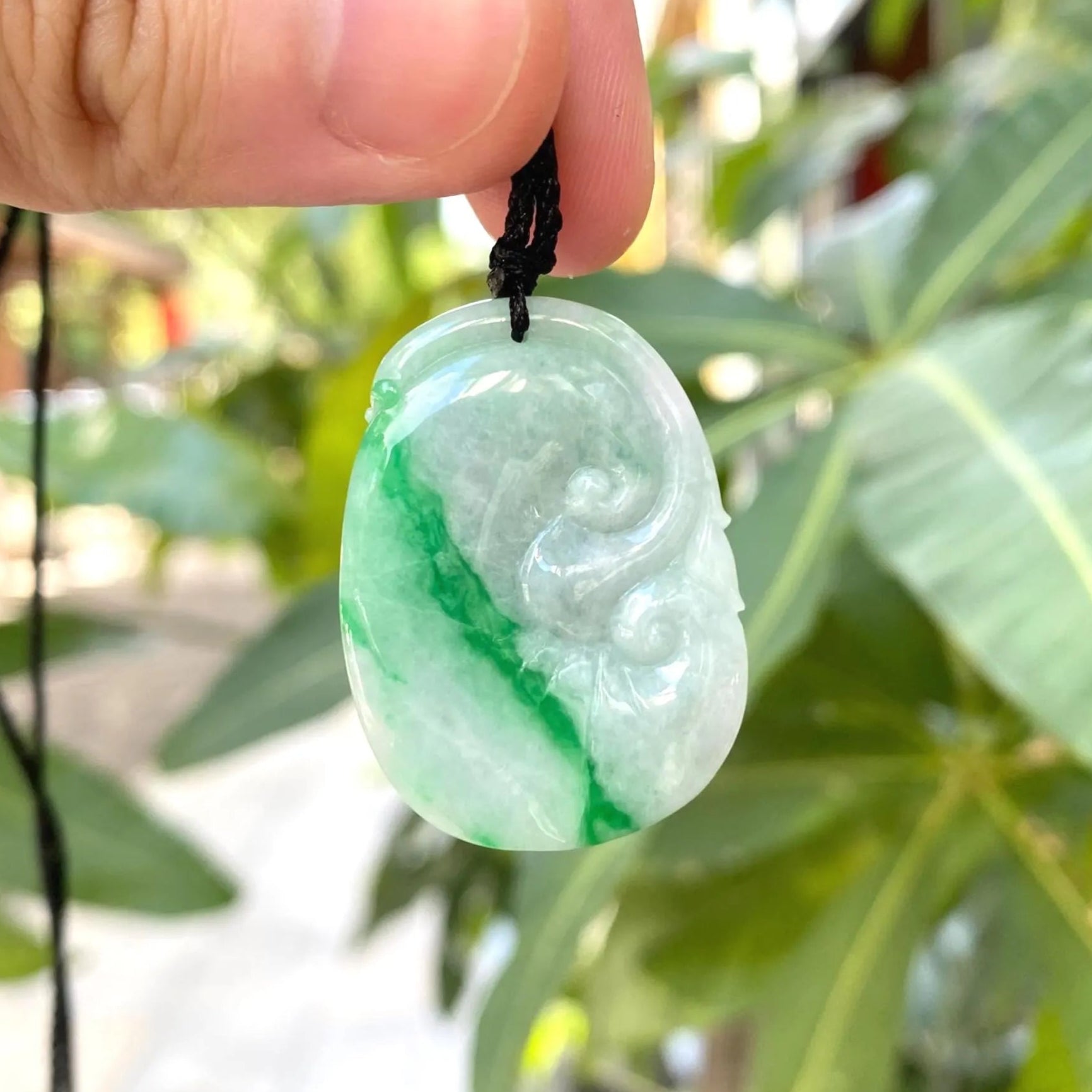 Green jade pendant held by a hand with a blurred green leafy background